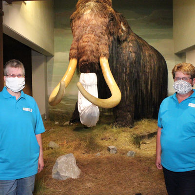 Bild vergr��ern: Museumsleiterin Dr. Corinna Nauck, Mitarbeiterin Regina Molsberger und Museumsmaskottchen Mammut Willy freuen sich, wieder Besucher im Museum Wilnsdorf begr��en zu d�rfen. Auch Willy tr�gt Schutzmaske - bei einem Mammut reicht ein "R�sselschutz", da Atmen, Spucken und nat�rlich Niesen �ber das gleiche Organ, den R�ssel, erfolgen.